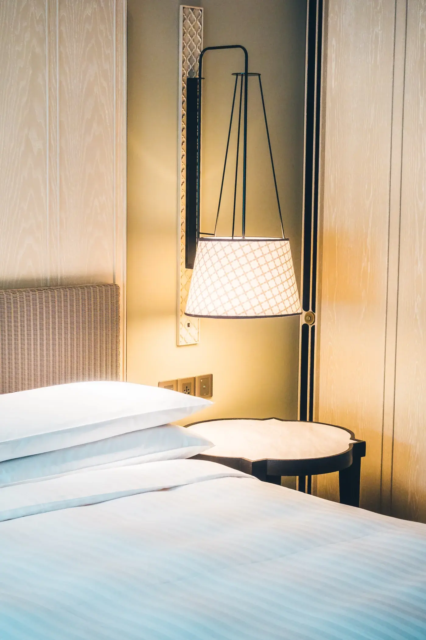 Letto bianco d’albergo accanto a lampada sospesa e tende scure
